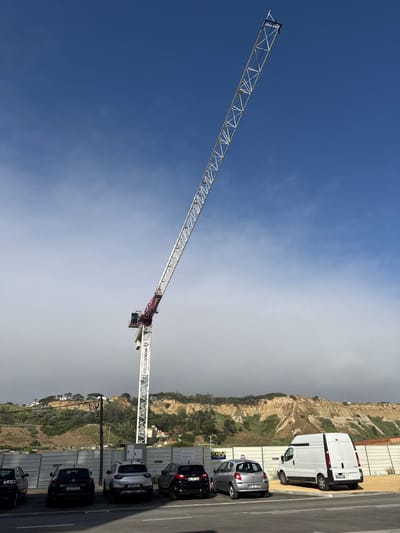 The image shows a construction site with a large tower crane extending diagonally across a blue and overcast sky. The crane is white with red accents and has the text "SUMAFUTURE" visible on its boom. In the background, a hilly landscape with vegetation and some buildings can be seen. There are several vehicles parked along a road next to a construction fence, including a white van and a few cars. The scene appears to be set during the daytime, with mixed weather conditions indicated by the varying sky colors. The overall impression is one of an active construction project in a coastal area. The location context is identified as Costa da Caparica, Portugal.