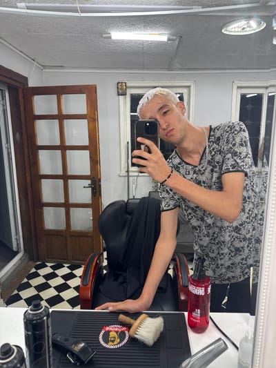 A young man with bleached blonde hair and a skull-patterned t-shirt takes a selfie in a barbershop. He is holding a smartphone and looking at the camera. To his right, a red bottle of hair product, a hair clipper, and a brush are on a white counter. Behind him, a black barber chair is positioned on a black and white checkered floor. A wooden door with glass panes is visible to the left of the chair. The barbershop appears to be located in Gurkovo, Bulgaria.