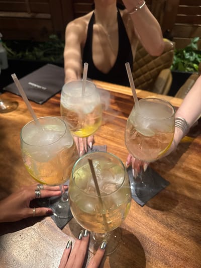 Four cocktails, likely gin and tonics, sit on a wooden table. The drinks are served in large stemmed glasses filled with ice, a straw, and a citrus slice. Each glass is emitting a cloud of vapor, possibly from dry ice. Hands with various rings and bracelets are poised near the glasses. One set of hands has mirrored chrome nails. Black coasters are visible under some of the glasses.