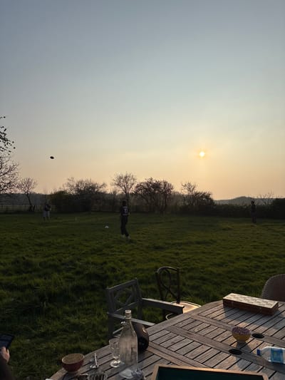 This image depicts an outdoor scene at sunset in Bû, France, characterized by a bright sun positioned in the upper right quadrant of the sky. The sky displays a gradient from orange-yellow near the horizon to a pale blue-grey overhead.
In the foreground, a slatted wooden outdoor dining table occupies the lower portion of the frame. On the table, from left to right, are: a red ceramic bowl, silverware, a clear glass bottle, two clear drinking glasses (one stemmed), an inverted dark bottle or container, a small yellow bowl with a pink interior, a white plastic container, and a rectangular wooden box with a patterned lid. A portion of a dark electronic device, possibly a tablet or phone, held by an unseen individual, is visible in the bottom-left corner. Two outdoor chairs are partially visible near the table, one wooden with a slatted back, and another with a curved back. A wicker-backed chair is visible on the far right.
The midground features a mown grassy field. Three individuals are present in the field. One person, positioned near the center-left, wears a dark shirt displaying the number "10" on the back, dark trousers, and light-colored footwear. A white soccer ball rests at their feet. Another individual, less distinct, is positioned further left in the field, wearing light-colored clothing. A third individual, also indistinct, is located on the far right of the field, wearing dark clothing. All three individuals are facing away from the camera. A dark, circular object, consistent with a frisbee or disc, is suspended in the air in the upper-left area of the field.
The background consists of a treeline composed of deciduous trees and bushes, some bare and some with early foliage, stretching across the horizon. A fence line is partially visible behind the individuals on the left. Faint outlines of distant hills or higher ground are discernable above the treeline on the right side of the image.