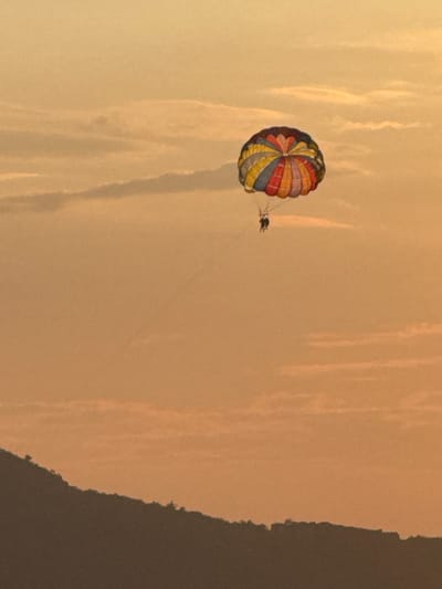 In Vrijesno, Montenegro, during what appears to be sunset or sunrise, two individuals are parasailing across a hazy, orange-toned sky. A brightly colored parachute, featuring segments of red, yellow, blue, and pink, is suspended high above a dark, silhouetted hillside that curves across the lower portion of the frame. The sky is filled with faint, horizontal cloud formations.