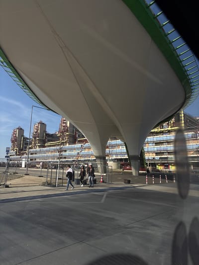 This image captures an exterior view of the Uniklinik RWTH Aachen in Aachen, Germany. A prominent, large, white tensile-fabric canopy, supported by two substantial pillars and featuring a green top edge, spans across the upper and central portions of the frame, serving as a modern architectural element, possibly a pedestrian bridge or covered walkway. Below this structure, on a paved sidewalk, four individuals are walking from left to right. They appear to be adults dressed in casual clothing.
In the background, the distinctive, multi-story building of the Uniklinik RWTH Aachen is clearly visible. It features a complex façade with numerous windows and a series of prominent, industrial-style towers on its roofline, characterized by exposed red-orange and yellow piping and structural elements.
The immediate foreground consists of dark asphalt pavement, possibly a road or parking area, with faint white markings. A temporary metal barrier fence runs along the left side of the sidewalk, separating it from an unpaved area. Red and white striped bollards and a single orange traffic cone are visible on the right side of the paved area. The sky above is clear blue with minimal cloud cover. Reflections in the bottom right corner suggest the photograph was taken from inside a vehicle.