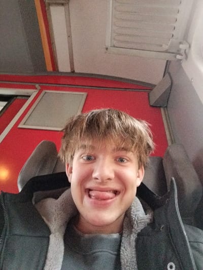 A young man with blond hair and blue eyes smiles, sticking his tongue out. He is seated on a train, facing the camera. The train's interior features red walls with framed windows and grey seating. He wears a grey sweater and a dark jacket with a furry collar. The lighting appears to be natural.