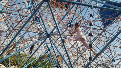 An upward-angled, wide shot captures a complex playground structure, possibly located in Kiryat Gat, Israel. The structure is composed of a network of blue steel pipes and brown rope netting. Two people are on the play structure: One person wears a light pink outfit. The other person wears a dark outfit. Patches of blue sky with scattered clouds are visible through the netting.