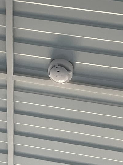 The image shows a white smoke detector mounted on a corrugated metal ceiling. The ceiling panels are grey with thin white lines along the corrugations. A vertical white metal beam is visible on the left side of the image, intersecting with the ceiling panels. The smoke detector has a slightly raised central section and appears to have some markings or buttons on its surface. The lighting suggests an indoor environment. There are no people or other objects of note in the image. The specific location in Usolye-Sibirskoye, Russia, is not determinable from the visual information provided.