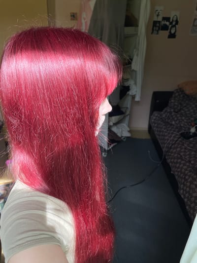 The image shows a close-up view of a person's bright red hair. The hair is long and appears to be styled with bangs that fall across the forehead. The person is wearing a light-colored, short-sleeved t-shirt. In the background, a bedroom is visible. There is a bed with patterned bedding, a dark-colored rug on the floor, and a closet or wardrobe with clothes hanging inside. On the wall, several black and white photographs are pinned up. The lighting suggests it is daytime, with sunlight casting a shadow on the person's shoulder and highlighting the hair. The scene is indoors, likely in a residential setting. The location of Солнечная Поляна, Russia is not evident from the image itself.
