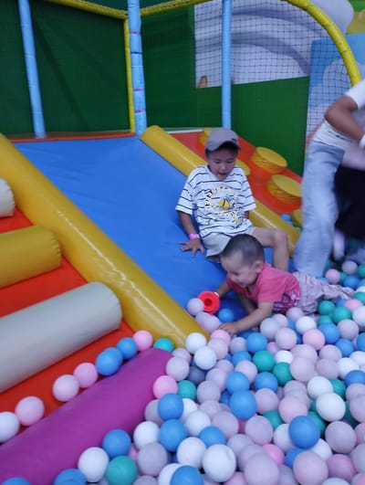 In a brightly colored indoor play area, possibly a soft play center, in Talas City, Kyrgyzstan, two young children are engaged in play. The environment features a blue slide with yellow padded rollers on either side, and a large area filled with soft, colorful balls in shades of pink, blue, white, and green.
A young boy, wearing a striped t-shirt and light-colored shorts, is seated on the blue slide. He is looking downwards with a slight smile. To his right and slightly below him, a younger child, a baby or toddler, lies amidst the ball pit. This child is wearing a pink t-shirt and appears to be reaching for a red toy, possibly a ring, with their right hand. The baby is surrounded by the colorful balls. In the background, parts of the play structure are visible, including blue and yellow padded poles and a green netting enclosure. The lighting suggests an indoor setting.