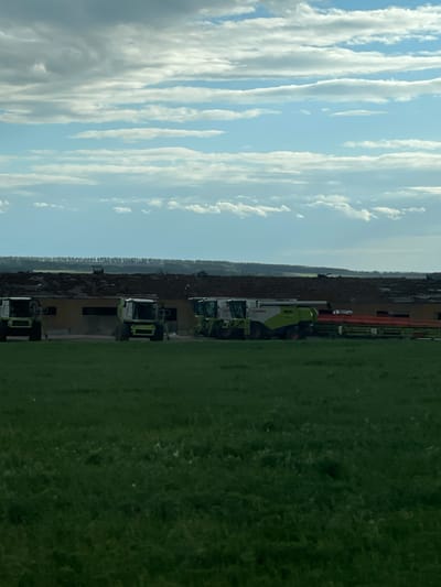The image shows a rural scene with a cloudy sky on the left and a green field on the right. In between, there are buildings and agricultural vehicles, including tractors and combine harvesters. The scene appears to be in an agricultural area. Specific details linking the image to Петрово, Russia are not discernible from the visual information.