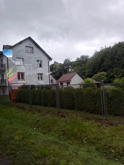 In Wilkowisko, Poland, on a cloudy day, a three-story residential building with a light gray facade is visible on the left. The building has several windows with white frames and dark shutters. A balcony with a dark railing is attached to the left side of the building. In front of the building, there is a dense hedge and a metal fence. To the right of the main building, there is a smaller, single-story house with a brown roof and white walls. Behind the houses, a dense forest of green trees covers the background. The foreground is filled with green grass and some weeds. A rainbow-like refraction is visible on the left side of the main building, possibly due to light passing through a window.