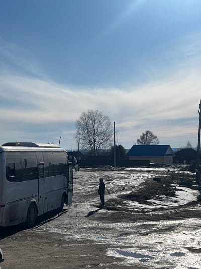 A horizontal, outdoor shot shows a partial view of a bus parked on a street next to a person walking on what appears to be a dirt or snow-covered road. The sky is light blue with light white clouds. In the background is a small house with a blue roof and bare trees. The road or pathway has some reflective patches, possibly from ice or water. The location is potentially Gory, Russia.