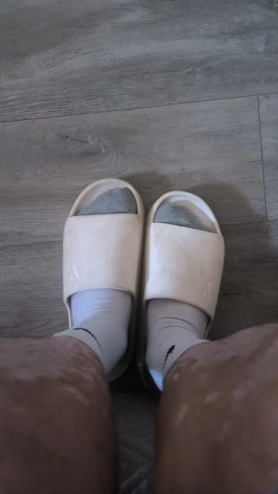 The image shows a close-up, top-down view of a person's feet wearing white slide sandals and white socks, standing on a grey wooden floor. The sandals have a simple, wide strap design. The floor appears to be made of laminate or wood planks with a visible grain pattern. The lighting suggests an indoor environment. There is no indication of the city of Kovrov, Russia, or any specific urban context in the image.