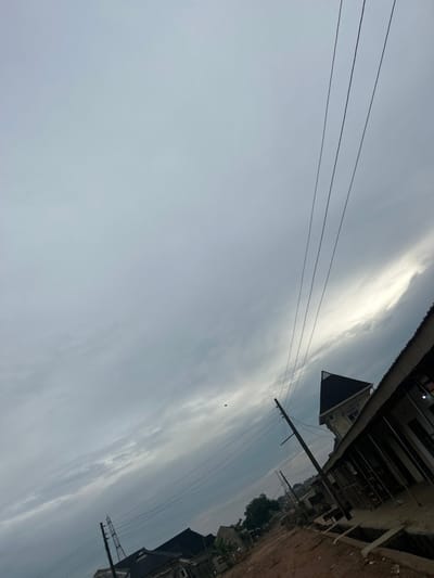 Overcast sky with a faint horizon line dividing the lower gray cloud cover from the upper lighter clouds. Power lines stretch across the sky. A building with a dark triangular roof is partially visible. Utility poles rise vertically. Residential buildings and trees line the ground in the background. A dirt path is visible at the upper right corner.