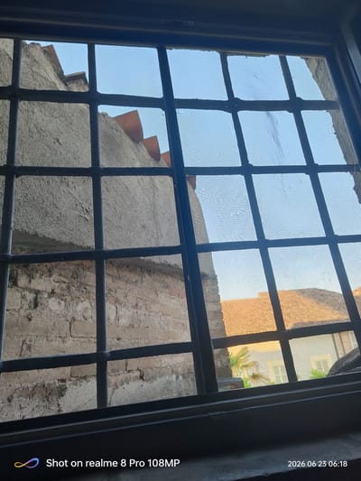 A view through a window with a black metal grille and a torn insect screen shows a section of a building's exterior. The building has textured, light-colored walls and what appears to be a brick layer beneath some sections. The roofline is visible with terracotta-colored elements. In the background, another building with a similarly colored roof and some foliage can be seen under a clear blue sky. The timestamp "2026 06 23 06:18" and "Shot on realme 8 Pro 108MP" are present in the lower right and left corners respectively. The location is identified as Foligno, Italy.