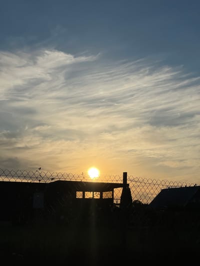 A sunset is visible behind a chain-link fence in Zaķumuiža, Latvia. The sun is a bright orb, positioned in the center of the frame, with rays of light extending outwards. The sky is a gradient of blue and yellow, with wispy clouds. In the foreground, the chain-link fence is silhouetted against the sky. Behind the fence, a dark building with two square windows is visible. The windows are illuminated by the setting sun. Trees and foliage are also visible in the background, silhouetted against the sky. The scene is tranquil and serene, with a sense of peace and quiet.