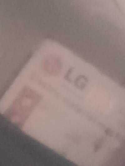 A highly out-of-focus image, captured in Elatan, Uzbekistan, depicts a light-colored, rectangular object. This object, possibly paper or packaging, features a blurred red circular logo positioned above the indistinct letters "LG" rendered in a dark, sans-serif font. Below the LG branding, several lines of completely illegible text are present. A reddish-brown rectangular shape with indiscernible markings occupies the lower left portion of the object. The bottom left corner of the object is partially obscured by a dark, soft-textured material, likely fabric. The background consists of a uniform, light grey surface. No individuals, specific actions, or interactions are discernible within the frame.