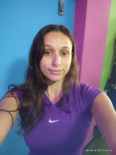 A female individual is depicted in a chest-up selfie taken indoors. She has long, dark brown hair, brown eyes, and fair skin with visible freckles on her face. She is wearing a purple V-neck short-sleeved top featuring a white Nike swoosh logo on the left chest. A thin, dark band is present on her left forearm. The background consists of a bright blue wall on the left, transitioning to a vertical pink column or wall section in the center-right, and a lime green wall section on the far right. A dark, metallic bracket is affixed to the blue wall in the upper left quadrant. The image bears a timestamp of "5 de mar. de 2023 11:53 a.m." The specific location within the city of Quilmes, Argentina, is not discernible from the visual evidence.