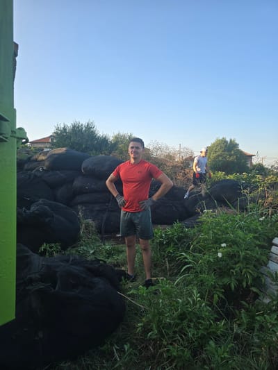 In this media, two men are visible in an outdoor setting, likely during the daytime given the bright sky. The scene appears to be a rural or semi-rural area, with abundant greenery and what looks like a pile of large black objects, possibly covered by tarps or netting, taking up a significant portion of the foreground. In the background, a building with a tiled roof and trees suggest a residential or agricultural area.
The man in the foreground is wearing a red t-shirt, dark shorts, and grey gloves. He is looking towards the camera with a smile and has his hands on his hips, adopting a relaxed pose. Behind him, and further into the scene, another man is wearing a light grey t-shirt, dark shorts, and a white cap. He is partially obscured by the foliage and the large black objects.
The activity seems to involve work or a task related to the large black items, perhaps related to agriculture or land management, given the outdoor setting and the presence of gardening gloves on the man in the foreground. The overall mood appears casual and positive, with the man in the foreground smiling. The lighting suggests it is either morning or late afternoon. There is no visible text in the image.