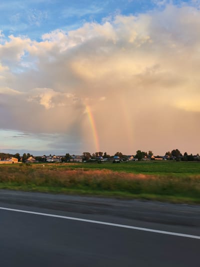The image captures a rural landscape with a prominent rainbow arcing across a sky filled with dramatic clouds, tinged with the warm hues of sunset. Below the sky, a small village is nestled amidst green fields, characterized by its cluster of houses and trees. In the foreground, a dark asphalt road with a white line runs horizontally, suggesting motion or a viewpoint from a moving vehicle. The overall scene is serene and picturesque, evoking a sense of natural beauty and tranquility in a rural Russian setting.