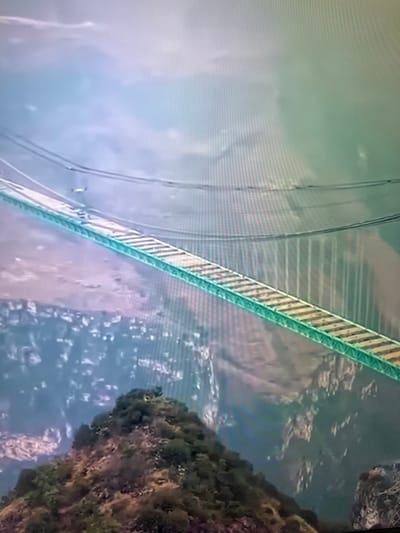 The image displays a long, narrow, suspended bridge with a green metal frame and a walkway made of wooden planks with yellow and white striped safety markings. The bridge spans across a vast, mountainous landscape with a hazy, possibly misty, sky. The perspective suggests an aerial view, looking down and across the bridge. In the foreground, a lush, green, and rocky hillside is visible. The bridge appears to be a significant structure in a remote, natural setting, with no obvious signs of urban development or specific landmarks that would tie it to the city of Vangaži, Latvia. The overall impression is one of a high-altitude crossing over rugged terrain.