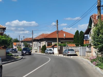 The media file captures a sunny day in a village, likely Krushovitsa, Bulgaria, given the context. The scene is a paved road curving through a residential area lined with houses featuring red tile roofs. Several cars are parked along the road and on driveways, including a dark SUV, a silver sedan, and a black hatchback. The architecture of the houses suggests a rural European setting, with some exhibiting traditional elements. A Bulgarian flag is visible, adding a national context to the location. The weather appears clear with some white clouds dotting a bright blue sky, indicating it's likely daytime, possibly late morning or early afternoon. The atmosphere is calm and serene, reflecting a typical village scene. There are no people actively engaged in any activity, and the focus is on the static elements of the environment. Visible text includes license plates on the cars. For example, one license plate reads "CT 4764 BK" and another appears to be "EN 7530 KA". The image composition, with the road leading into the distance, creates a sense of depth and invites the viewer to imagine further exploration of the village. The presence of trees and some flowering plants adds a touch of greenery and color to the scene.