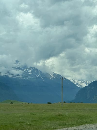 The image shows a vast green grassy plain stretching towards a range of imposing mountains. The sky is heavily overcast with thick, grey clouds, and some of the mountain peaks are capped with snow and partially obscured by clouds. A few power lines are visible in the foreground, running horizontally across the frame. A solitary utility pole with wires stands in the middle ground, slightly to the right of center. There are no people or animals visible in the scene. The overall impression is one of a wild, natural landscape under a moody sky. The location is identified as Nizhnyaya Teberda, Russia.