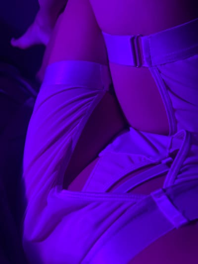 A close-up, low-angle shot shows a person's lower torso clad in white lingerie. The lingerie features adjustable straps and what appears to be a mesh fabric panel. The lighting is predominantly purple. The location in Heenatigala Junction, Sri Lanka is not discernible from the image.