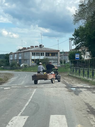 The image shows a rural scene in the city of Маминское, Russia. Two men are riding on a small agricultural vehicle with a trailer attached, moving down a paved road. The vehicle appears to be a tractor or a similar utility vehicle. The trailer is carrying several items, including what looks like sacks or boxes, and some bottles. The road is marked with white lines and shows signs of wear and tear, with some patches of dirt and puddles.
In the background, there are residential buildings, typical of a village or a small town. One building is a two-story apartment block with multiple windows and balconies, and chimneys on the roof. Another building, further in the background, appears to be a smaller house. A bus stop sign is visible on the right side of the road. The environment is characterized by green vegetation, trees, and a cloudy sky. The overall impression is a snapshot of daily life in a rural Russian setting.