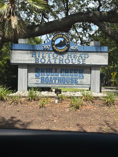 This image shows a wooden sign with blue lettering at the entrance to "HILTON HEAD BOATHOUSE" and "SKULL CREEK BOATHOUSE" located at 397 and 405 on Hilton Head Island, South Carolina, United States of America. The sign is weathered and has a nautical theme, featuring a silhouette of a boat within a circular emblem. It is surrounded by trees and foliage, and the ground in front of the sign is covered with mulch and small plants. The sign appears to be situated along a road, as indicated by a yellow road sign visible in the background. There are no people or animals visible in the image.