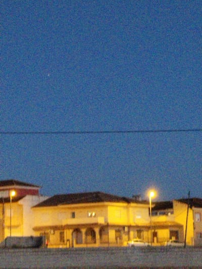 A row of houses sits under a deep blue sky, illuminated by streetlights. The houses are low and wide, with dark roofs and lit windows. Cars are parked in front of the houses. A thin dark line stretches across the sky above the houses. The sky is textured and grainy.