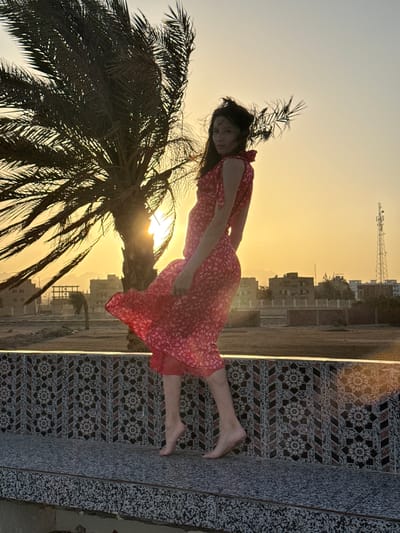 The image captures a woman posing outdoors during sunset or sunrise, set against an urban and natural backdrop. The main subject is a woman with dark hair, wearing a flowing red dress adorned with a white floral or patterned design. She is barefoot and appears to be leaning back or standing against a decorative tiled wall or barrier on the right side of the frame. Her head is tilted towards the camera, and she looks slightly upwards with a serene expression. Her arms are gently bent, resting near her waist.

On the left side, a large palm tree with dark, silhouetted fronds occupies the foreground. The sun, a bright glow, is visible through the palm fronds, casting a warm, golden light that dominates the sky. The sky transitions from a vibrant yellow-orange near the horizon to a lighter, paler hue higher up, characteristic of twilight.

In the mid-ground and background, an urban landscape is visible beneath the glowing sky. Buildings of various sizes, likely residential, are spread across the horizon, along with a distinct, tall communication tower. The patterned tiled surface next to the woman features intricate geometric designs, resembling star-like or snowflake patterns within squares, bordered by stripes. These tiles are predominantly dark brown, black, and white. There is also another section of wall or ground with a speckled, granite-like texture.

The overall scene conveys a sense of peaceful beauty, with the woman striking a composed pose amidst the warm, golden glow of the sun. There is no visible text in the image. The presence of the palm tree and the architectural style are consistent with the provided location context of Safaga, Egypt.