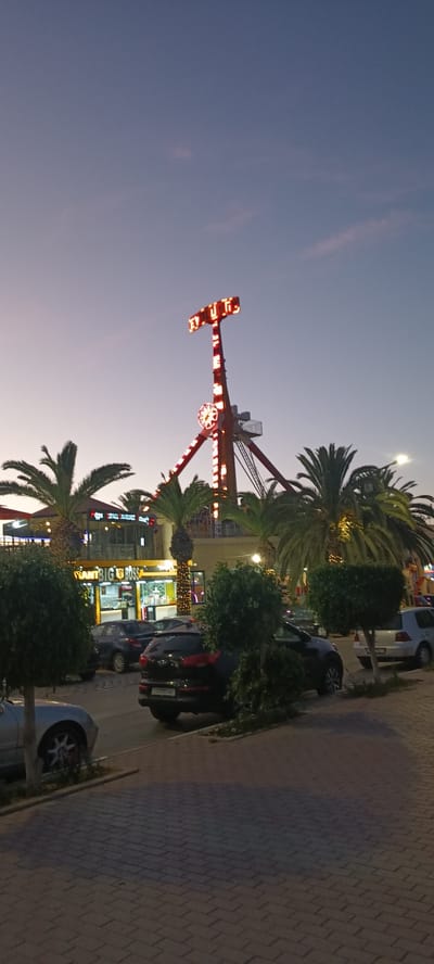 This is a vertical, full-shot photograph taken at dusk in Hammam Sousse, Tunisia, depicting an amusement park setting. In the background, a tall amusement ride with red and white lights is prominently displayed against a gradient twilight sky. The word "TITUS" is illuminated in red letters at the top of the ride. Palm trees frame the ride and buildings below, suggesting a warm climate. In the middle ground, a building with yellow signage reads "Restaurant BIG BOSS" and "SPACE AMOUR". Several cars are parked in front of the restaurant. In the foreground, meticulously pruned green bushes and trees line a paved walkway. The overall impression is one of a lively entertainment district at the close of day.