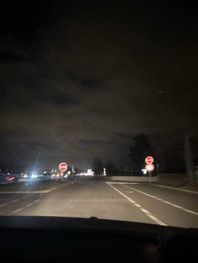 A nighttime view from a car driving on a road with clear lane markings. Several illuminated road signs, including stop signs, are visible along the side of the road. Other cars' headlights pierce through the dark background. The sky appears cloudy or foggy. The scene conveys a sense of motion and darkness.
