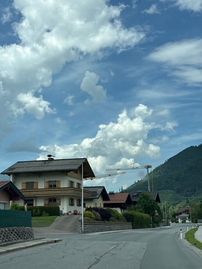 The image depicts a view of a street in Bruck an der Großglocknerstraße, Austria. The foreground shows a paved road with white lane markings and reflective bollards. To the left, a residential property is visible with a dark green fence and a low stone wall. Further back, a multi-story house with a dark tiled roof, wooden shutters, and a balcony is prominent. Adjacent to this house are other residential buildings with pitched roofs and well-maintained gardens. In the background, a steep, tree-covered mountain rises, and two construction cranes are visible against a bright blue sky with scattered white clouds. The overall scene suggests a residential area undergoing some development or construction, situated within a mountainous landscape.