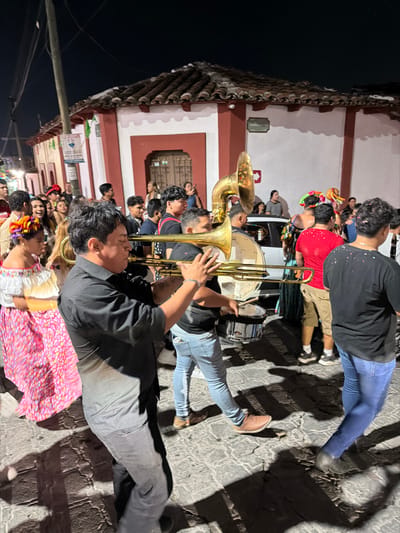 A group of musicians are playing brass instruments, including a trombone, tuba, and drums, while standing on a cobblestone street at night. The musicians are mostly men, and they are dressed in casual clothes. Some people are wearing traditional Mexican clothing. The background shows a building with a red and white facade, illuminated in the dark setting.