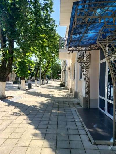 The image depicts a sunny day on a sidewalk lined with trees and a building in Labinsk, Russia. The sidewalk is paved with square tiles and features tree grates around the bases of the trees. The trees are mature with lush green foliage, and their trunks are whitewashed at the bottom. Along the right side of the sidewalk is a building with a light-colored facade. An awning made of blue translucent material and ornate metalwork extends over the entrance of the building. The awning is supported by decorative metal brackets. The windows and doors of the building are visible. The perspective of the shot is looking down the sidewalk, with the building on the right and the trees and a street on the left. The sunlight casts strong shadows across the sidewalk. There are no people or vehicles clearly visible in the image, suggesting a quiet moment.
