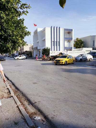 In Raoued, Tunisia, on a sunny day, a yellow sedan, possibly a Peugeot 308 based on the license plate, is parked on the right side of a street. The license plate reads "199 TUN 6331". Next to it, a silver sedan is parked. The street is paved and shows signs of wear, with a prominent curb on the left side of the frame, bordered by dry leaves and debris.
In the background, a modern white building with blue vertical accents is visible. A Tunisian flag flies from the top of the building. A few people are scattered around the scene, including one individual in a pink dress walking away from the camera on the sidewalk near the building. A person's arm and shoulder, wearing a pink shirt, are visible on the far left of the image, suggesting they are walking towards the intersection. Further down the street, more cars and pedestrians are present, along with trees and other buildings. The sky is clear and blue. A black pickup truck loaded with produce is parked on the left side of the street in front of a building with Arabic writing on its facade. The overall scene depicts a street in a Tunisian city with typical urban elements.