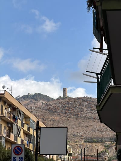 This image captures a vibrant daytime scene in Maddaloni, Italy, showcasing a blend of urban architecture and natural mountainous landscape. The sky, a clear blue with scattered white clouds, dominates the upper portion of the frame.
In the foreground, on the left, part of a building is visible, featuring a dark green metal awning or pergola with clothesline supports extending outwards. Below this, some dark green foliage adds a touch of nature. On the right, a multi-story, light-colored (possibly beige or yellow) residential building occupies the lower-right section. This building features multiple balconies, windows, and numerous air conditioning units, with a satellite dish mounted on its roof.
The midground and background are dominated by a large, rugged, brown rocky mountain or hill, covered in sparse, dry vegetation, indicating an arid environment. A distinct, weathered stone tower or ruin is perched prominently on the left slope of this mountain, adding historical character to the landscape. Further to the right on the mountain, a smaller, light-colored building is nestled among the rocks.
Above the building on the right, a large, rectangular blank billboard or sign stands out. Below it, attached to a pole, is a roadside sign. This sign prominently displays a red circular "No Stopping/Standing" symbol (blue background with a red border and a diagonal line). Below this, a blue square contains a white "P" indicating parking, alongside a white symbol for disabled parking.
The scene is illuminated by bright daylight, suggesting midday or early afternoon. No people or specific activities are clearly visible, presenting a calm, static snapshot of everyday life in this Italian town nestled against a dramatic natural backdrop.