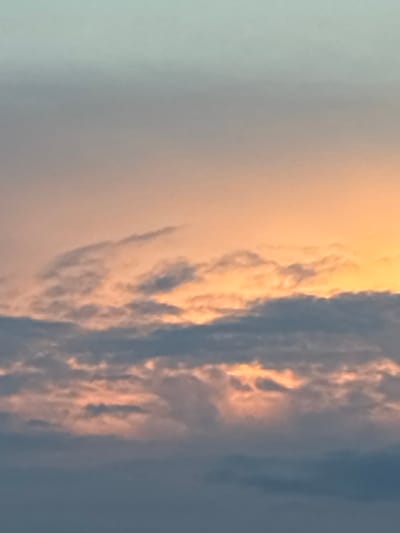 This is a photograph of a sky filled with clouds at sunset or sunrise. The sky transitions from a pale blue at the top to a warm orange hue lower down, illuminated by the setting or rising sun. Several layers of clouds are visible. The top layer consists of wispy, thin clouds tinged with orange. Below this, a thicker layer of darker, greyish-blue clouds is present, with some sections catching the light and appearing with orange highlights. The very bottom of the image shows a denser, darker cloud layer. The scene is entirely natural and does not contain any identifiable landmarks or structures from Toruń, Poland, or any people or objects.