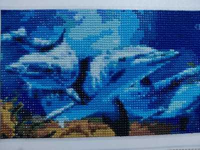 This is a close-up, horizontal shot of a diamond painting depicting an underwater scene. The artwork is composed of numerous small, faceted beads in various shades of blue, white, black, and brown, arranged in a grid to form an image. The primary subjects are several white sharks swimming in deep blue water. Lighter blue and white beads create a sense of light filtering from above and the frothy texture of water. Darker blue beads suggest depth. In the foreground, a textured seabed is depicted with brown, tan, and dark green beads, possibly representing sand and seaweed or coral formations. There are no people visible, and the activity is a serene underwater environment. The lack of specific time-of-day or weather cues, typical for an underwater scene, and the absence of discernible text reinforce the focus on the marine life and its habitat. The overall impression is one of a dynamic yet peaceful aquatic setting.