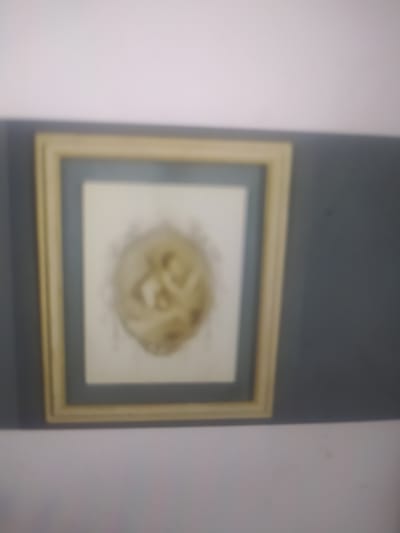 The image shows a framed picture on a wall. The frame is light-colored, possibly gold or beige, with a darker mat inside. The picture itself appears to be a circular image with figures within it. The background wall is painted a light color, and there is a dark band running horizontally behind the frame. The overall image is slightly out of focus. There is no discernible information about the location in Pshekhskaya, Russia.
