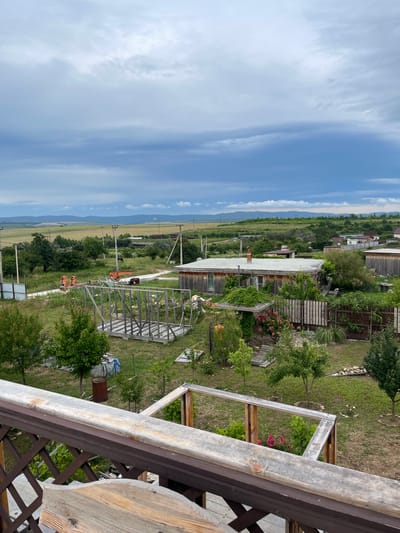 The image depicts a rural landscape with a construction site and residential buildings in Gay-Kodzor, Russia. In the foreground, a wooden railing and part of a wooden table are visible, suggesting a vantage point from a balcony or porch. The mid-ground shows an unfinished wooden structure, likely a shed or a small house, surrounded by a grassy yard with scattered trees and bushes. Several construction materials and tools, including orange planks and a rusty barrel, are present. Further back, a long, single-story wooden house with a chimney and a red-tiled roof is situated, partially covered with ivy. Wooden fences delineate the property boundaries. In the background, a dirt road winds through the area, with more residential buildings and power lines. The scene extends to a wide expanse of fields, hills, and a cloudy sky, indicating an overcast day. There are several individuals in the distance near the construction area, wearing orange safety vests, suggesting ongoing work. The overall impression is that of a working rural property undergoing construction or expansion.