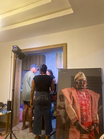 A photograph taken indoors depicts a film set in what seems to be a private residence. The photo is oriented vertically, showing a corner of a room with beige-painted walls and a doorway. On the wall is a framed portrait of an elderly man in traditional Nigerian attire.

In the doorway, a film crew appears to be working. At least three people are visible; one is wearing a black shirt and blue jeans, facing towards the interior room, another wearing a light-colored shirt and a baseball cap. Camera lights and equipment surround them. The interior room is tiled and contains wooden cabinetry.

Given the mention of Ado-Ekiti, Nigeria, the portrait subject is likely a local figure.
