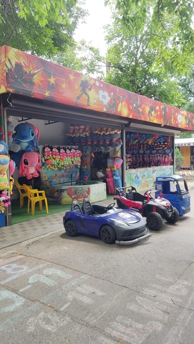 The image depicts an outdoor amusement game stall with several plush toys hanging behind a counter. In front of the stall, there are three small, rideable electric toy cars for children. A blue sports car is in the foreground, followed by a red and black ATV, and then a blue truck. The stall itself has a vibrant awning with comic book-style graphics. Behind the counter, various plush toys, including large Stitch and Angel characters, are displayed. There's also a small water game with toy ducks and a person sitting at a table inside the stall. The ground in front of the stall is paved, with chalk drawings on it. The scene appears to be set in a park or recreational area, possibly in the city of Novaya Adygeya, Russia, given the context provided. The overall atmosphere suggests a fun and engaging environment for children.