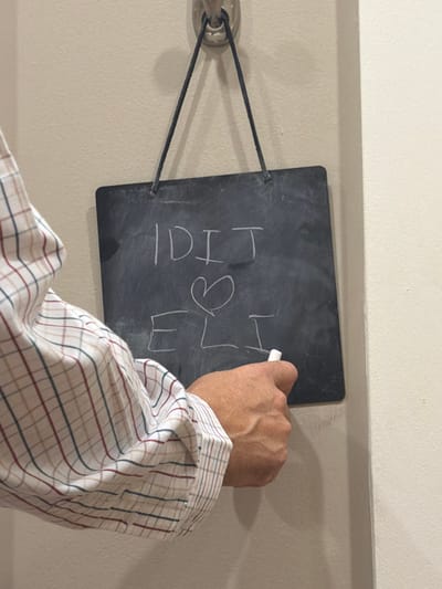 A close-up, indoor image captures a person's right arm and hand actively writing on a small, dark-colored chalkboard. The individual wears a long-sleeved plaid shirt with white, red, blue, and brown lines. The right hand holds a white piece of chalk, positioned to complete the letter "I" as part of the name "ELI" on the lower portion of the chalkboard. Above "ELI," a small heart symbol is drawn, and on the top line, the name "IDIT" is written in capital letters. The rectangular chalkboard, featuring rounded corners, is suspended from a silver-colored hook on a light-colored wall (off-white or light beige) by a black cord. A vertical structural edge is visible on the far right of the frame. The precise location within the city of Hackensack, United States, is not discernible from the visual content.