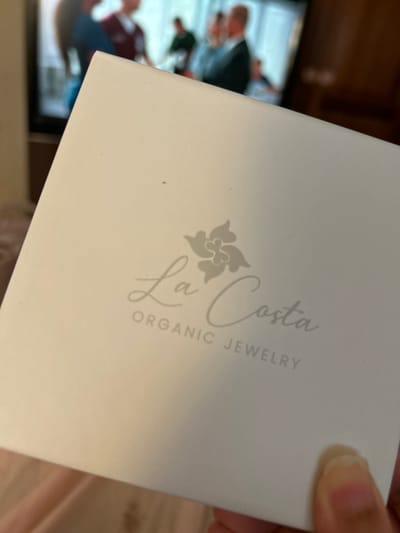 The image shows a white box with the words "La Costa Organic Jewelry" printed on it in grey. The box is being held by a hand. The background is blurry, showing a television screen with a group of people in it. The coordinates provided place the location in the Colonial Zone, Santo Domingo, Dominican Republic.