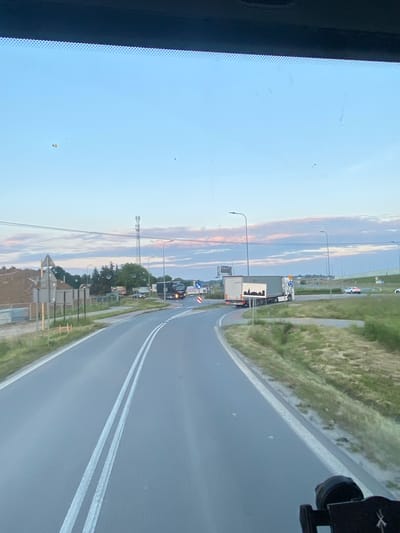 The image shows a view from a vehicle driving on a paved road with double white lines in the city of Podłęże, Poland. The sky is a pale blue with wispy pink and white clouds. On the right side of the road, there is a grassy embankment and a highway barrier. In the distance, streetlights and a cell tower are visible. On the left side of the road, there is a construction site with a large pile of dirt and some fencing. A few vehicles, including trucks and a car, are on the road and surrounding areas. The image appears to have been taken during the late afternoon or early evening.