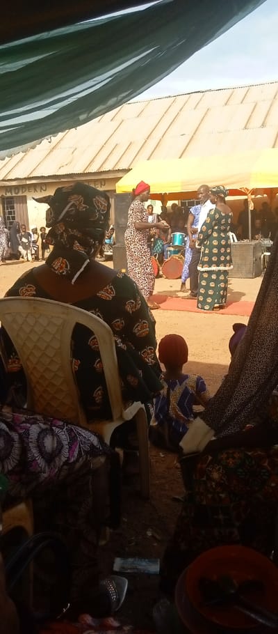 The scene depicts an outdoor gathering in Kasuwan Magani, Nigeria, characterized by a reddish-brown dirt ground and partially shaded by a dark green sheer canopy in the foreground. In the immediate foreground, a person wearing a dark green patterned garment with crown motifs and a matching headwrap sits facing away in a white plastic chair. To their right, a small child in a blue and yellow patterned outfit with a reddish head covering sits on the ground, also facing away. An orange bowl with dark contents and a spoon, alongside a piece of light blue paper, are visible on the ground at the bottom right.
In the midground, three adults stand on a red rug. A woman in a light brown leopard-print dress and red headwrap stands on the left, holding a small, light object in her hands while facing right. A man in a light blue and white patterned shirt stands centrally, facing slightly left towards the first woman. A second woman, wearing a dark green patterned dress with crown motifs and a dark headwrap, stands on the right, facing left towards the man. Behind these figures, under a yellow canopy, numerous other individuals, including adults and children, are congregated. A group of children in white tops stands to the far left. Musical instruments, including a light blue drum set and two large brown traditional drums, are present, along with sound system equipment (speakers/amplifiers) positioned on either side of the standing adults.
The background features a building with a light-colored corrugated metal roof, bearing a sign that reads "MODERN QUEEN." The sky above is partially visible, appearing light and cloudy.