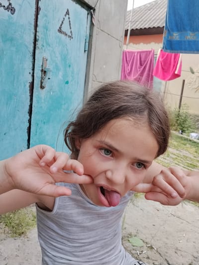 A young girl with brown hair and blue eyes makes a silly face, sticking out her tongue and pulling at her cheeks with her fingers. She is wearing a gray tank top. The background shows a turquoise-colored metal door with a latch and some markings, likely a shed or garage door. To the right, a clothesline hangs with colorful towels and clothing, including blue Nike towels. In the distance, there's a hint of a building with a corrugated roof and some greenery. The scene appears to be outdoors on a bright day, given the lighting. The girl's expression suggests playfulness and a candid moment. The location is identified as Vulcănești, Moldova.