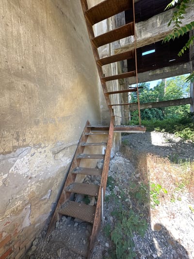An external view shows a rusty metal staircase leading up along a weathered, concrete wall. The wall has visible graffiti and peeling paint. Beyond the staircase, through an opening in a derelict building structure, a lush green canopy of trees is visible, suggesting an outdoor or semi-outdoor environment. The ground surrounding the staircase is covered with rubble and dry vegetation. The overall scene depicts an abandoned or decaying industrial site within the city of Rosen, Bulgaria.