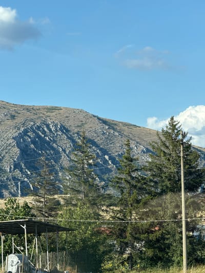 The image depicts a mountainous landscape with a clear blue sky and scattered clouds. In the foreground, there are several evergreen trees, some power lines, and a utility pole. A partial view of a covered structure with supporting poles is visible on the left, along with some debris. The background features a large, rocky mountain with sparse vegetation. The scene appears to be in a rural or undeveloped area, possibly on the outskirts of the city of Dragoman, Bulgaria.
